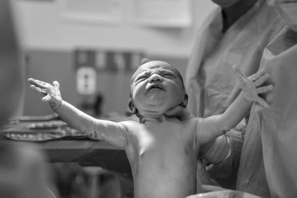 A newborn baby is being held by medical staff in a hospital setting. The baby is extending its arms and appears to have just been delivered. The scene is captured in black and white, highlighting the textures and emotions of the moment.