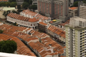 Aerial view of a tightly packed urban area featuring rows of buildings with red-tiled roofs. The architectural style appears to be a mix of older, possibly colonial buildings and modern high-rise structures. The area is surrounded by greenery and includes a variety of dense residential and commercial buildings. The image displays a somewhat compressed and miniature-like effect, likely due to the photography technique used.