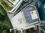 An aerial view of an urban landscape showcasing a building with a rooftop solar panel installation. The scene is surrounded by lush greenery, curved pathways, and additional buildings, suggesting a blend of nature and modern architecture.