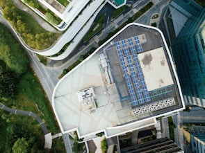 An aerial view of an urban landscape showcasing a building with a rooftop solar panel installation. The scene is surrounded by lush greenery, curved pathways, and additional buildings, suggesting a blend of nature and modern architecture.