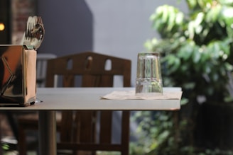A wooden table set in an outdoor or garden setting, featuring a metal napkin holder with utensils and a glass placed upside down on top of paper napkins. The background includes lush green foliage.