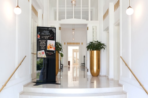 A bright, elegant hallway with white walls and golden accents. Two spherical pendant lights are hanging from the ceiling. A black sandwich board displays a menu with various food items. Large gold planters with green plants are placed on either side of the hallway, enhancing the upscale atmosphere.