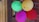 A cluster of colorful balloons with fun and cartoonish facial drawings hangs against a wooden and brick wall. The balloons are green, yellow, red, pink, and blue, each featuring different illustrated faces.