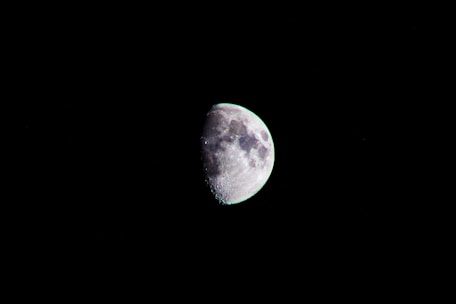 A detailed view of the moon in a partly illuminated crescent phase against a stark black background. The lunar surface features a mix of light and dark areas indicating craters and maria.