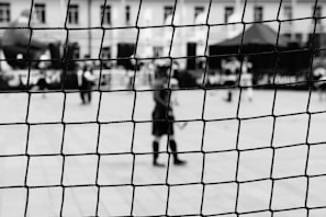 A black and white photograph captures a sports net in sharp focus in the foreground, while blurred figures and structures are visible in the background. The setting appears to be an outdoor sports area or event.