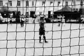 A black and white photograph captures a sports net in sharp focus in the foreground, while blurred figures and structures are visible in the background. The setting appears to be an outdoor sports area or event.