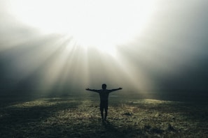 A person stands with arms outstretched in an open field, surrounded by mist with rays of sunlight streaming down from above, creating a serene and contemplative atmosphere.