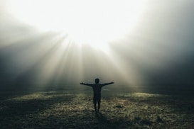 A person stands with arms outstretched in an open field, surrounded by mist with rays of sunlight streaming down from above, creating a serene and contemplative atmosphere.