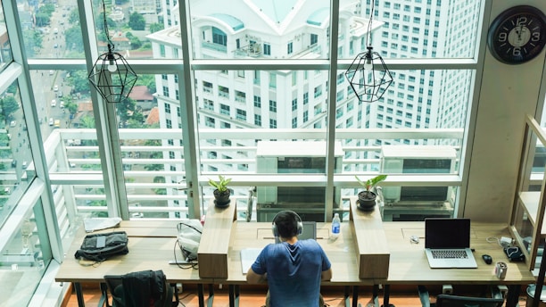 A person wearing headphones sits at a desk in a spacious, modern office with large windows overlooking a cityscape of tall buildings. The workspace has multiple laptops, potted plants, and a clock on the wall. Two geometric pendant lights hang from the ceiling, and there are additional seats with backpacks and bags.