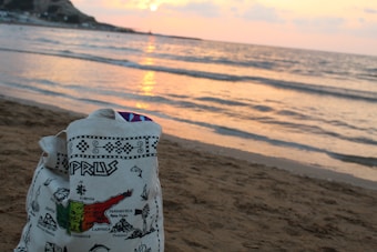 A canvas bag with a map and names of locations printed on it rests on a sandy beach. The ocean waves gently meet the shore at sunset, casting a warm glow on the water, while hills are visible in the distance.
