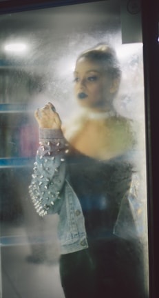 A person with dark makeup gazes through a fogged-up glass, wearing a denim jacket adorned with spikes. The atmosphere is dimly lit, creating a mysterious and introspective vibe.