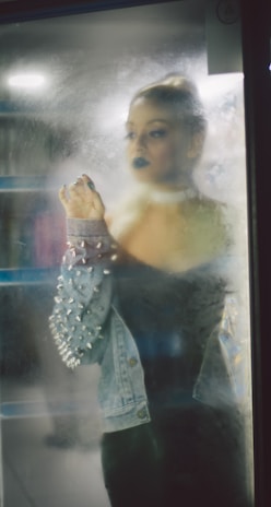 A person with dark makeup gazes through a fogged-up glass, wearing a denim jacket adorned with spikes. The atmosphere is dimly lit, creating a mysterious and introspective vibe.