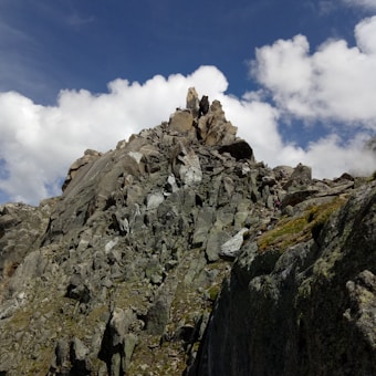A rugged, rocky mountain peak with jagged formations under a bright blue sky scattered with white, fluffy clouds. The surface of the mountain is a mixture of gray and brown stones with patches of moss. A small figure is visible climbing up the slope, adding a sense of scale to the scene.