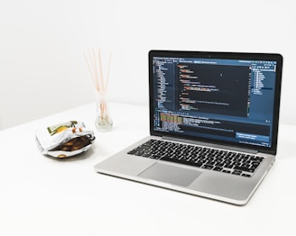 A laptop displaying code on its screen sits on a white desk. Next to the laptop, an open bag of snacks and a glass container with reed diffusers are visible. The setting appears to be minimalistic and organized, suggesting a workspace environment.