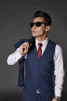 A man wearing a blue waistcoat with matching trousers, sunglasses, and a white shirt paired with a red tie. He holds the matching jacket over his shoulder casually, with a confident stance.
