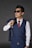 A man wearing a blue waistcoat with matching trousers, sunglasses, and a white shirt paired with a red tie. He holds the matching jacket over his shoulder casually, with a confident stance.