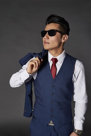A man wearing a blue waistcoat with matching trousers, sunglasses, and a white shirt paired with a red tie. He holds the matching jacket over his shoulder casually, with a confident stance.