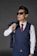 A man wearing a blue waistcoat with matching trousers, sunglasses, and a white shirt paired with a red tie. He holds the matching jacket over his shoulder casually, with a confident stance.