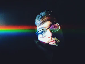 A person with glasses is partially illuminated by a vibrant, horizontal rainbow light across their face against a dark background. The lighting creates a dramatic and colorful contrast, highlighting the person's facial features.