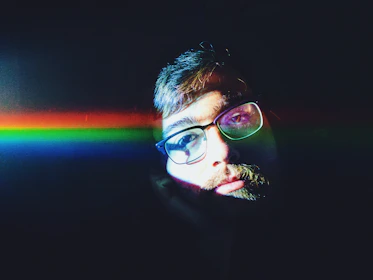 A person with glasses is partially illuminated by a vibrant, horizontal rainbow light across their face against a dark background. The lighting creates a dramatic and colorful contrast, highlighting the person's facial features.