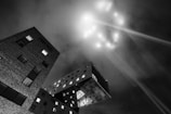 A stark black and white photograph of an architectural scene, featuring modern buildings with geometric shapes. The angle provides a dramatic perspective looking upward at a cantilevered structure. In the night sky, there are beams of light and glowing spots creating a mysterious and futuristic effect.