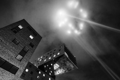 A stark black and white photograph of an architectural scene, featuring modern buildings with geometric shapes. The angle provides a dramatic perspective looking upward at a cantilevered structure. In the night sky, there are beams of light and glowing spots creating a mysterious and futuristic effect.