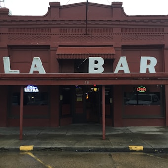A rustic red brick building with an old-fashioned sign displaying large, spaced letters spelling 'LA BAR'. There are neon signs in the window indicating the presence of a bar, including a 'Michelob Ultra' advertisement and an 'OPEN' sign. The entrance is situated under a covered awning with a number above the door.