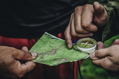 Hands are holding a topographic map and a compass. One hand is pointing at a specific location on the map, indicating navigation or orientation. The map is detailed with green shades, representing terrain or elevation.