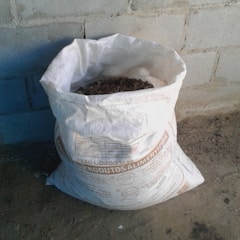 A white bag filled with organic material is placed on a rough concrete floor against a brick wall. The bag has text printed on it, likely indicating its contents and weight.