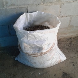 A white bag filled with organic material is placed on a rough concrete floor against a brick wall. The bag has text printed on it, likely indicating its contents and weight.