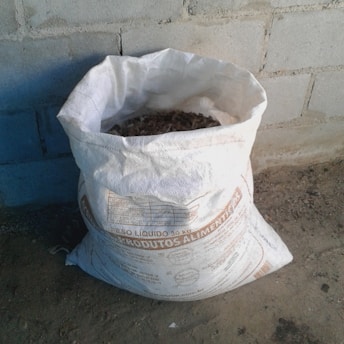 A white bag filled with organic material is placed on a rough concrete floor against a brick wall. The bag has text printed on it, likely indicating its contents and weight.