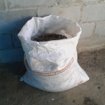 A white bag filled with organic material is placed on a rough concrete floor against a brick wall. The bag has text printed on it, likely indicating its contents and weight.