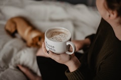 woman holding white ceramic teacup sitting on white blanket near short-coated tan dog