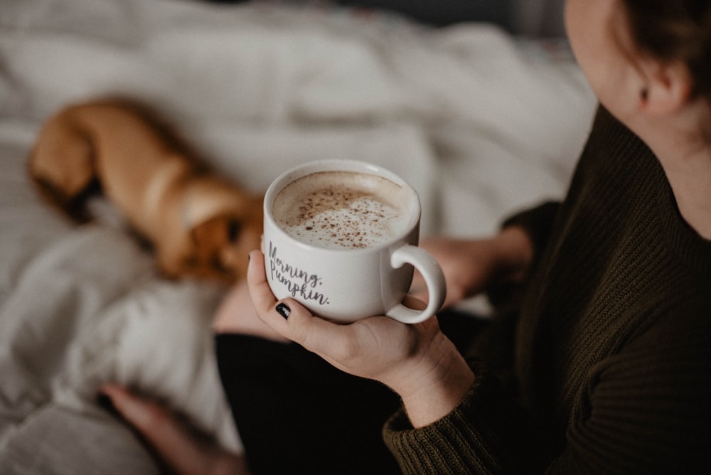 woman holding white ceramic teacup sitting on white blanket near short-coated tan dog