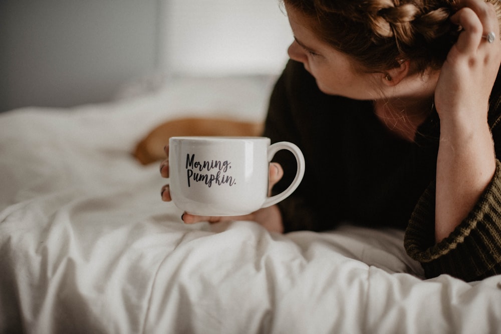 woman holding white mug while lying on bed