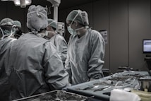 Medical professionals in surgical attire are gathered around an operating table in a hospital operating room. The room is equipped with medical instruments and equipment, including a computer monitor, surgical tools on a tray, and overhead lights.