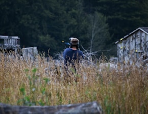 A person wearing protective gear appears to be in a field or woodland area surrounded by tall grass. Old wooden structures with visible yellow paint splatters suggest a paintball field environment.