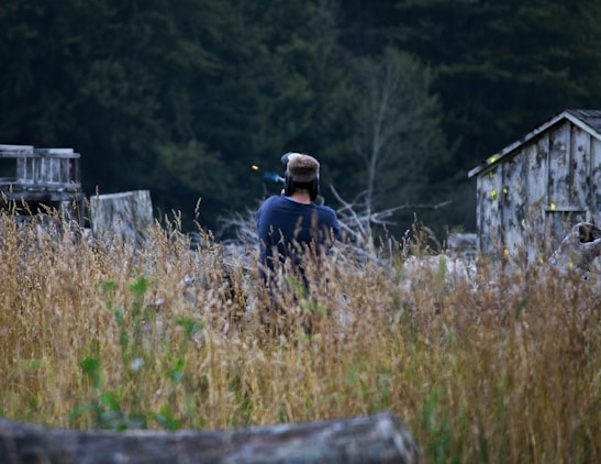 A person wearing protective gear appears to be in a field or woodland area surrounded by tall grass. Old wooden structures with visible yellow paint splatters suggest a paintball field environment.