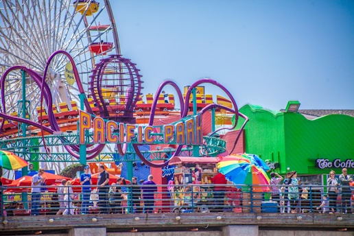 A vibrant amusement park scene featuring colorful signage for 'Pacific Park.' A large Ferris wheel is visible in the background along with roller coaster tracks looping overhead. People are walking around enjoying the attractions, and vividly colored umbrellas are scattered throughout.