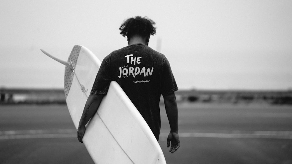 grayscale photo of man walking holding surfboard