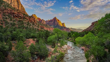 A picturesque landscape featuring towering red rock cliffs illuminated by sunlight, lush green trees along a winding river, and a clear blue sky with scattered clouds. People are seen enjoying the water in the river, adding a sense of activity to the serene natural setting.