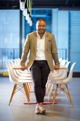 A person is leaning back on a row of white, modern chairs in a well-lit room with a polished concrete floor. The individual is dressed in a beige blazer, white shirt, and dark jeans, complemented by colorful striped socks and brown shoes. Overhead lighting and a blurred cityscape background add to the modern office setting.
