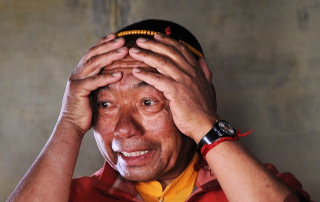 A person holding their head with both hands appears distressed or overwhelmed. They wear a red outer garment over a yellow shirt and have a red string bracelet on their wrist. A watch is visible on the left wrist, and the individual has a shaved head adorned with small beads.