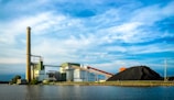 A large industrial facility is situated by the water, featuring a tall smokestack and several industrial buildings. A large pile of coal is visible, with conveyor belts running through the facility. The sky is partly cloudy, creating a backdrop of blue and white hues.