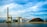 A large industrial facility is situated by the water, featuring a tall smokestack and several industrial buildings. A large pile of coal is visible, with conveyor belts running through the facility. The sky is partly cloudy, creating a backdrop of blue and white hues.