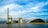 A large industrial facility is situated by the water, featuring a tall smokestack and several industrial buildings. A large pile of coal is visible, with conveyor belts running through the facility. The sky is partly cloudy, creating a backdrop of blue and white hues.