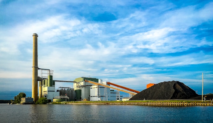 A large industrial facility is situated by the water, featuring a tall smokestack and several industrial buildings. A large pile of coal is visible, with conveyor belts running through the facility. The sky is partly cloudy, creating a backdrop of blue and white hues.
