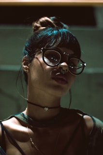 A person with large round glasses and a septum piercing, wearing a choker with a small heart charm. The lighting is dim and casts dramatic shadows on the face, with darker tones emphasizing the features.