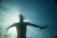 A silhouette of a person swimming underwater, with sunlight streaming through the water from above, creating a radiant effect. The person's arms are spread wide, holding an object in one hand.