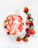 A bowl of smoothie garnished with fresh sliced strawberries, oats, coconut flakes, and small white and red flowers. An elegant spoon rests in the bowl, and additional strawberries and flowers decorate the surrounding surface.